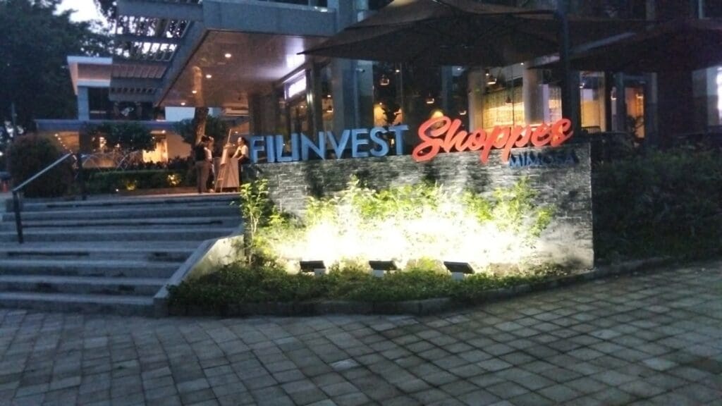 Acrylic outdoor signage for Filinvest Shoppee Mimosa is illuminated in front of a modern building with glass walls, plants, and outdoor steps at dusk in the Philippines, sign maker Philippines by M&G Global Ads.
