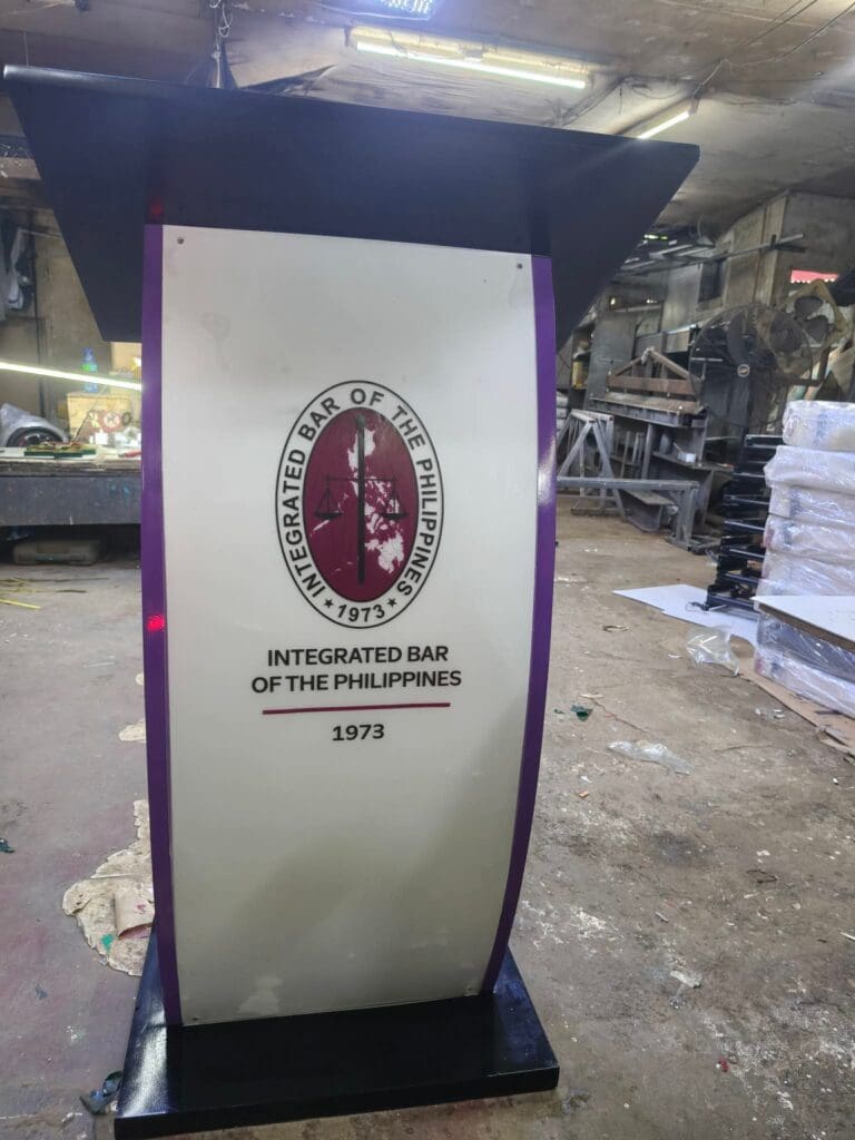 Acrylic podium signage featuring the Integrated Bar of the Philippines logo and “1973” on the front, placed in a cluttered workshop or storage area in the Philippines, sign maker Philippines by M&G Global Ads.