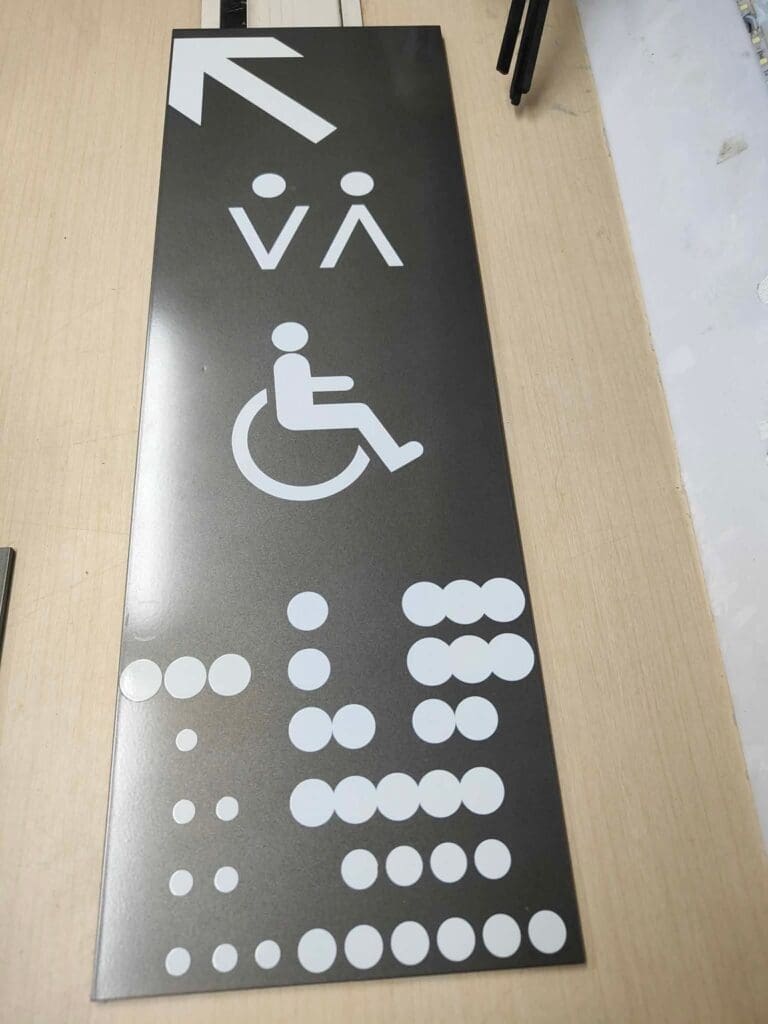 Acrylic wayfinding sign with male, female, wheelchair icons, braille; light wood background; sign maker Philippines by M&G Global Ads.