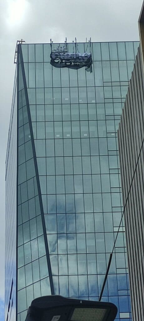 Building signage with workers on suspended platform cleaning a modern glass facade, Metro Manila, sign maker Philippines by M&G Global Ads.