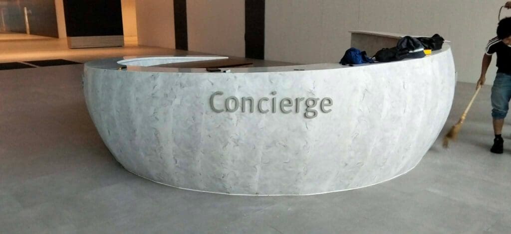 Curved, modern white concierge desk with stainless steel indoor signage displaying the word “Concierge” on the front, cleaning supplies on the counter, and a person sweeping nearby. Sign maker Philippines by M&G Global Ads. Location: Philippines.