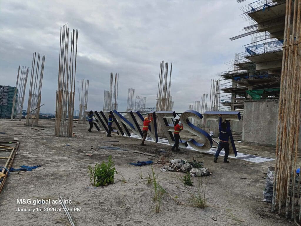 Five workers carry stainless steel building signage letters spelling INVEST on rooftop construction, Metro Manila, sign maker Philippines by M&G Global Ads.