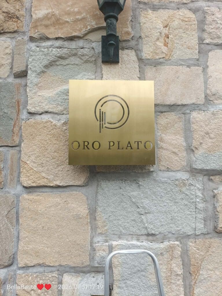 Gold brass building signage for ORO PLATO with circular logo, wall-mounted on a stone surface in the Philippines, small ladder visible below; sign maker Philippines by M&G Global Ads.