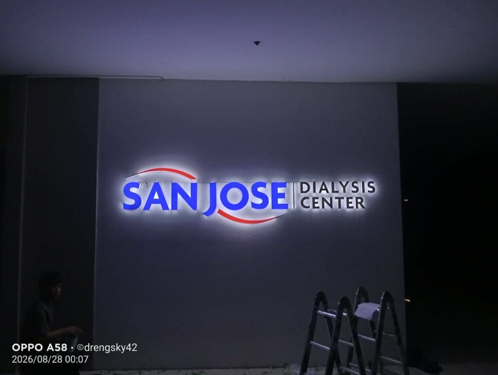 Illuminated LED building signage for SAN JOSE DIALYSIS CENTER on a wall at night, with a person partially visible in the lower left and a ladder on the right, Metro Manila, sign maker Philippines by M&G Global Ads.