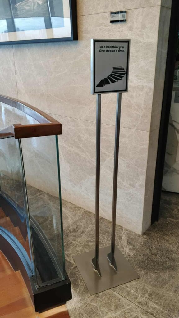 Indoor wayfinding sign on metal stands in a building lobby, marble floors, glass railings. Sign maker Philippines by M&G Global Ads.