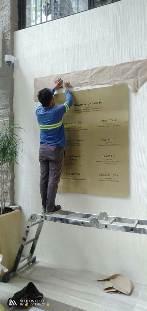 Man unveils engraved brass indoor plaque signage on white wall, Metro Manila, by sign maker Philippines M&G Global Ads.