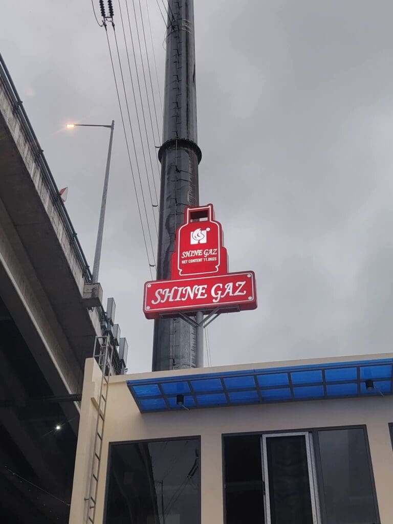 Outdoor pylon acrylic signage for SHINE GAZ featuring a red, bottle-shaped design, mounted on a tall pole near a building and an overpass under gray cloudy skies in Metro Manila by sign maker Philippines, M&G Global Ads.