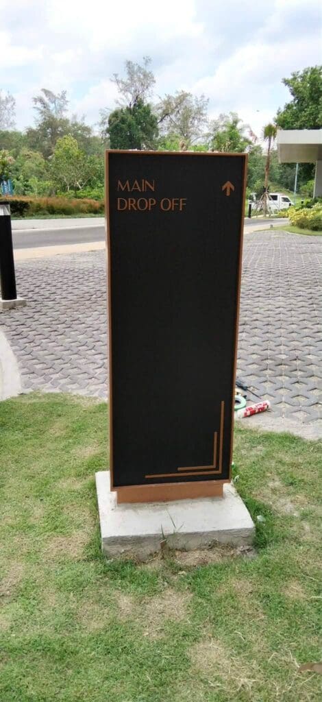 Outdoor wayfinding sign, possibly made of acrylic or aluminum, with white "MAIN DROP OFF" text and an upward arrow, positioned on a grassy area next to a paved road in the Philippines. Sign maker Philippines by M&G Global Ads.