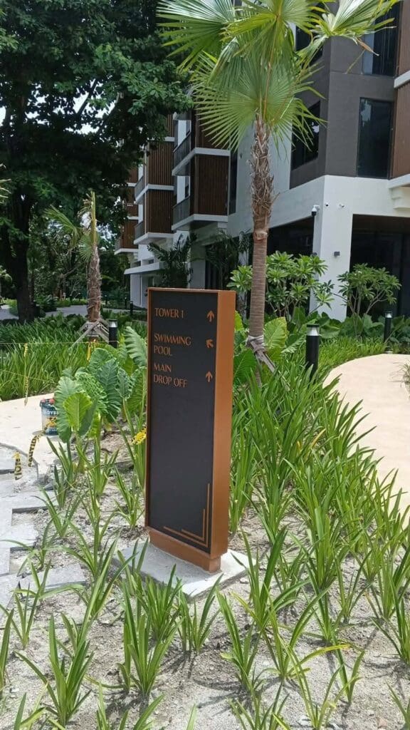 Outdoor wayfinding signage in a landscaped garden directs visitors to Tower 1, Swimming Pool, and Main Drop Off, with modern buildings and palm trees in the background. Location: Philippines. Sign maker Philippines by M&G Global Ads.