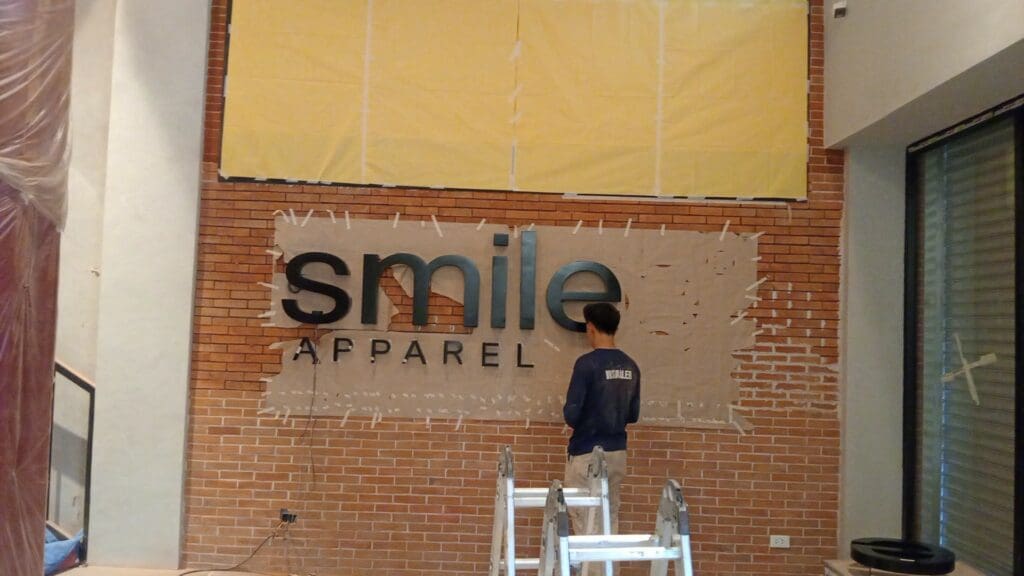 Person installs SMILE APPAREL building sign, acrylic signage on brick wall, Metro Manila sign maker Philippines by M&G Global Ads.