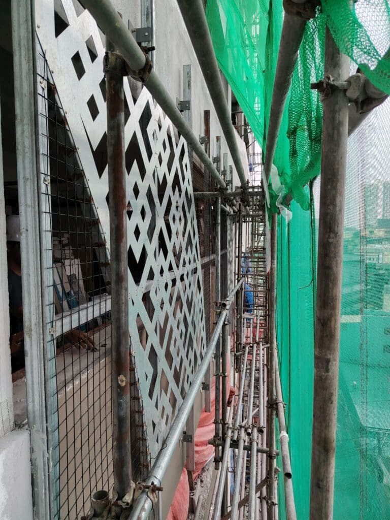 Outdoor building signage installation with metal scaffolding, patterned metal panels, and green safety netting along a facade, overlooking a cityscape in Metro Manila. Construction materials visible in the background. Stainless steel signage by sign maker Philippines by M&G Global Ads.