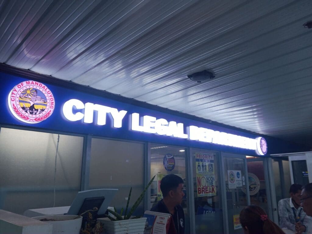 Indoor acrylic room signage for CITY LEGAL DEPARTMENT with the City of Mandaluyong seal, installed above glass doors in an office hallway featuring priority and no noon break signs. Client: City of Mandaluyong, Location: Metro Manila. Sign maker Philippines by M&G Global Ads.
