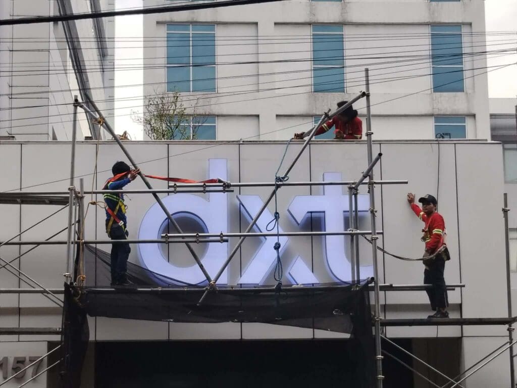 Three workers on scaffolding install or maintain a large illuminated LED building sign labeled “dxt” on a building exterior; safety harnesses and ropes are visible. A multi-story white building is in the background. Sign maker Philippines by M&G Global Ads. Location: Metro Manila.