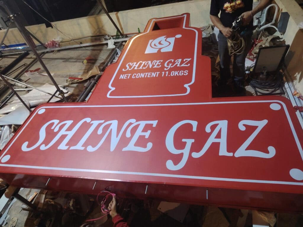 Outdoor acrylic signage in the shape of a red gas cylinder with white text reading “SHINE GAZ NET CONTENT 11.0KGS,” located in Metro Manila, with two people partially visible working nearby amid tools and other materials, sign maker Philippines by M&G Global Ads.