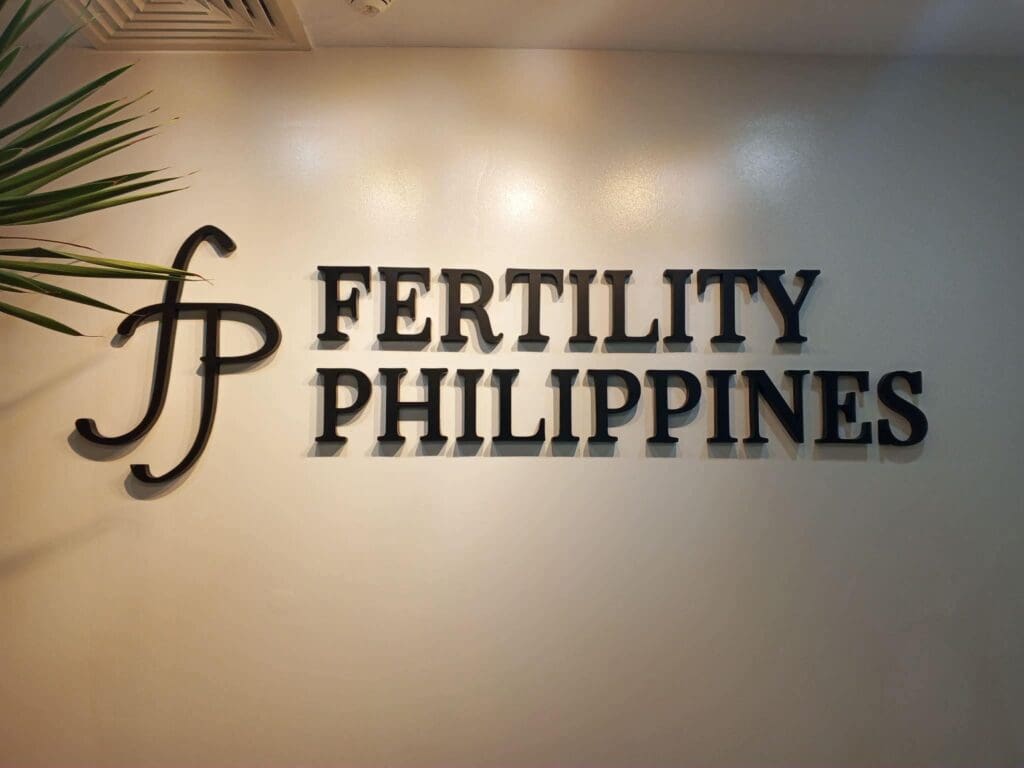Acrylic indoor wall sign with a stylized FP logo and bold black text reading FERTILITY PHILIPPINES, mounted on a light-colored wall with a plant visible on the left side of the image, for Fertility Philippines in the Philippines; sign maker Philippines by M&G Global Ads.