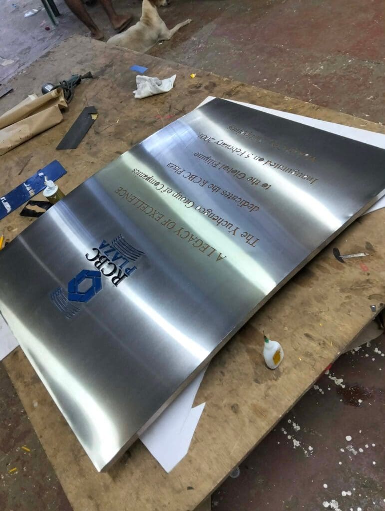 Stainless steel plaque signage with blue and gold text and the PRC logo by sign maker Philippines, displayed on a workbench with tools, glue, and papers at a workshop in Metro Manila; a dog is lying on the floor in the background. By M&G Global Ads.