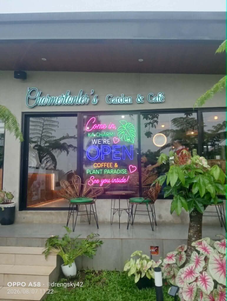 A café exterior in the Philippines featuring outdoor LED and neon signage displaying “Come in,” “WERE OPEN,” and “Coffee & Plant Paradise.” The main building sign above the entrance reads “Charmentonlers Garden & Café.” Visible elements include numerous plants, outdoor chairs, and large windows revealing a cozy interior. Sign maker Philippines by M&G Global Ads.