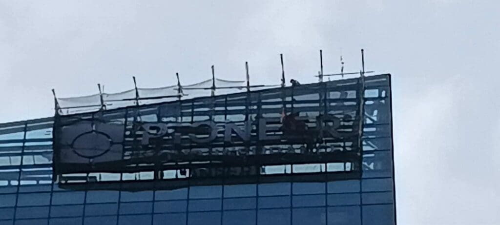 Sign maker Philippines installing outdoor building signage on glass facade, Metro Manila. Overcast sky. By M&G Global Ads.
