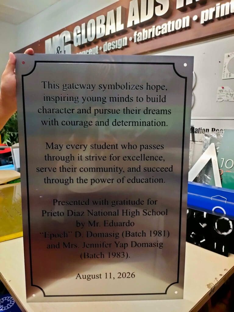 Stainless steel engraved plaque for Prieto Diaz National High School, featuring an inscription about hope, courage, excellence, and gratitude, dated August 11, 2026; shown held by a person in a workspace; sign maker Philippines by M&G Global Ads.