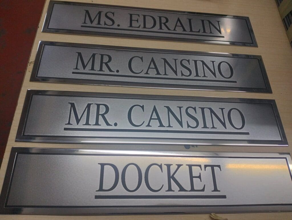 Stainless steel engraved room nameplates on a table, labeled MS. EDRALIN, MR. CANSINO, MR. CANSINO, and DOCKET; indoor wayfinding signage by sign maker Philippines in Metro Manila by M&G Global Ads.