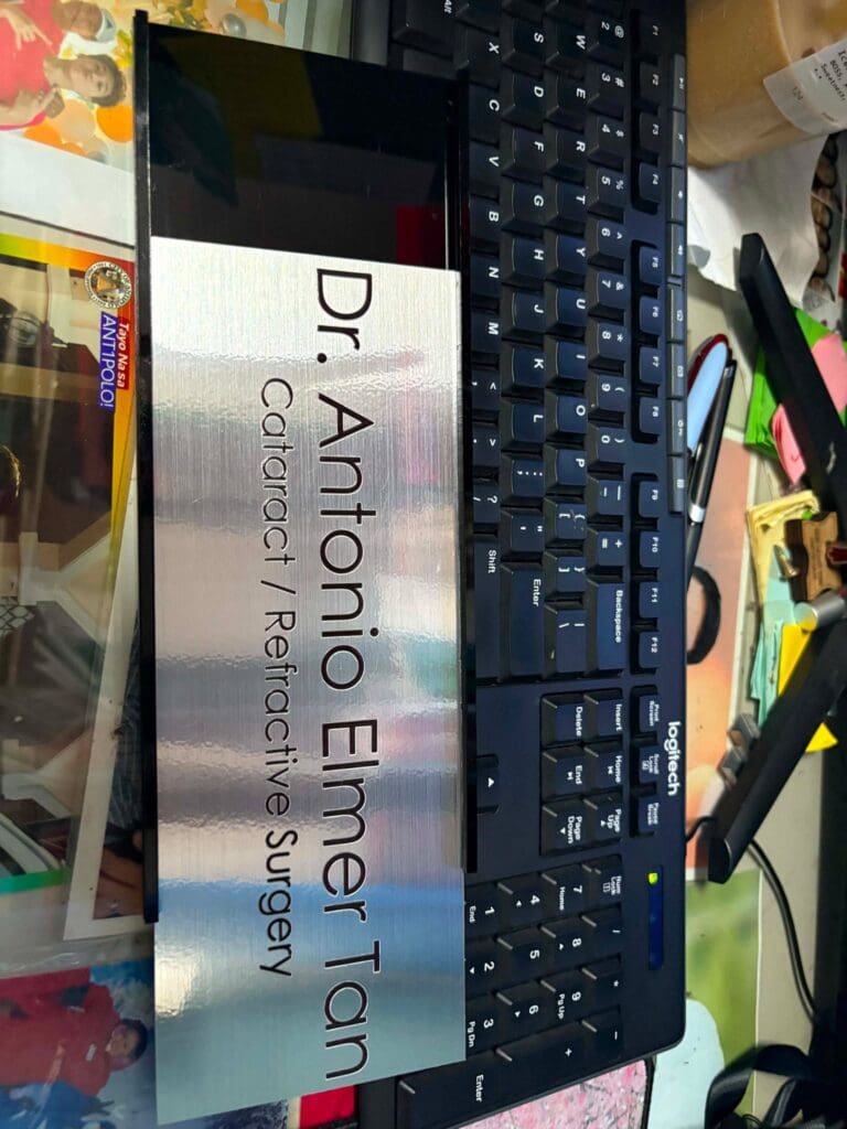 Stainless steel indoor nameplate signage for Dr. Antonio Elmer Tan, Cataract / Refractive Surgery, shown on a cluttered desk with a black keyboard and colorful papers, sign maker Philippines by M&G Global Ads.