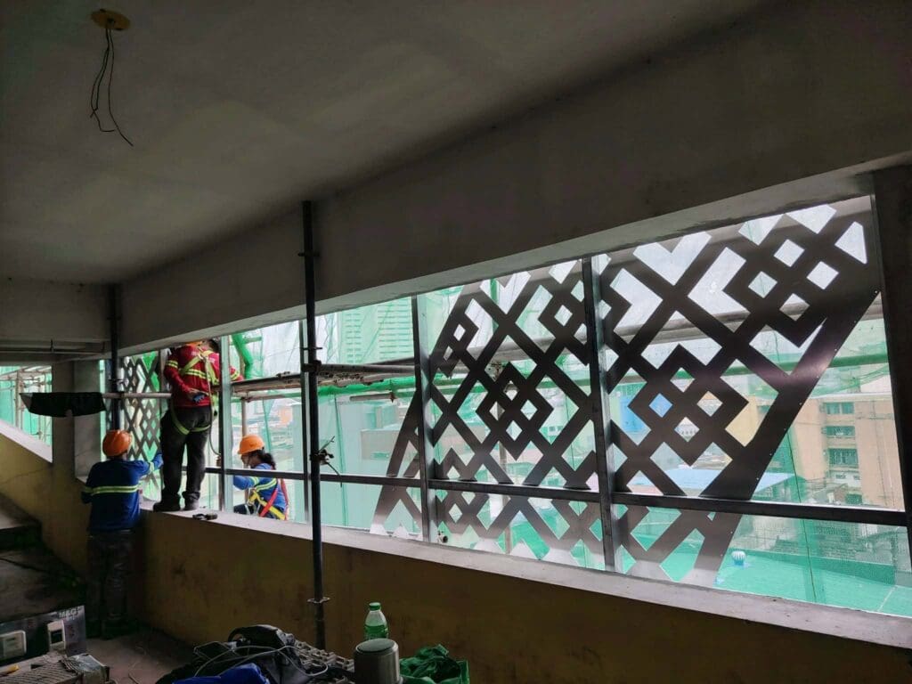 Three construction workers in safety gear install patterned metal building panels near large windows overlooking city buildings, Metro Manila, sign maker Philippines by M&G Global Ads.