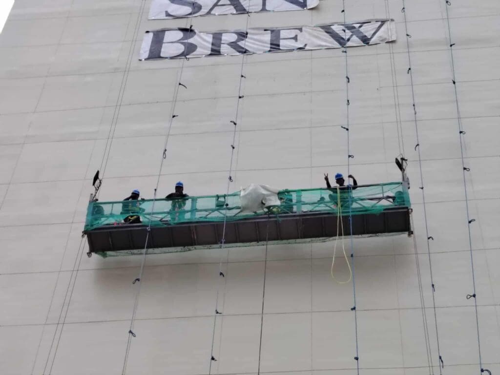 Three workers install an outdoor building signage banner (SAN BREW) on scaffold, sign maker Philippines by M&G Global Ads.