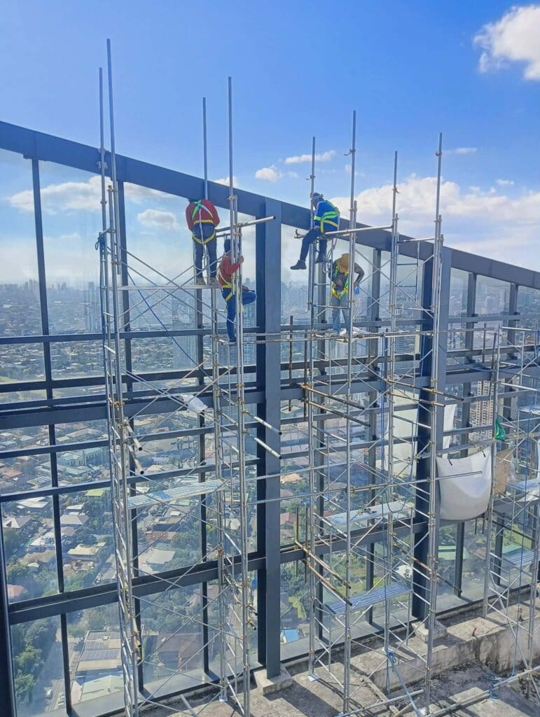 Three workers on scaffolding installing outdoor building signage, high-rise, city view, Metro Manila sign maker Philippines by M&G Global Ads.
