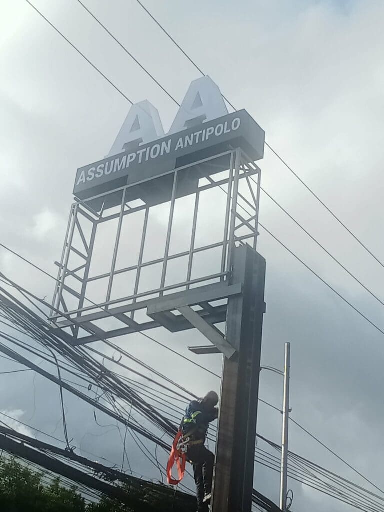 Worker installs ASSUMPTION ANTIPOLO outdoor building signage on tall metal frame, Antipolo, sign maker Philippines by M&G Global Ads.
