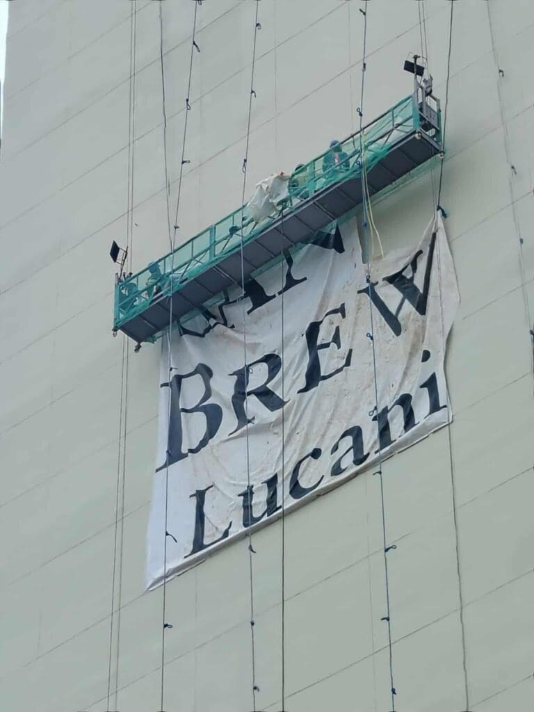Worker on suspended platform installs outdoor building banner signage for BREW Lucani, Metro Manila. Sign maker Philippines by M&G Global Ads.
