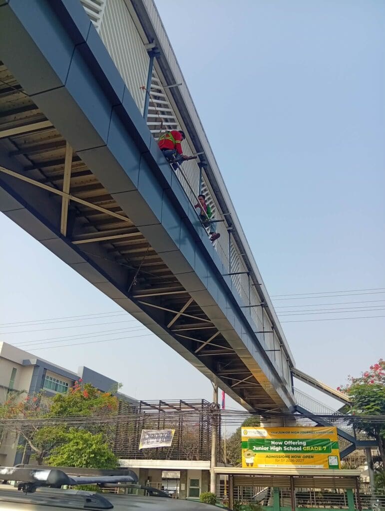 Workers clean a pedestrian overpass; yellow acrylic banner signage for junior high. Metro Manila sign maker Philippines by M&G Global Ads.