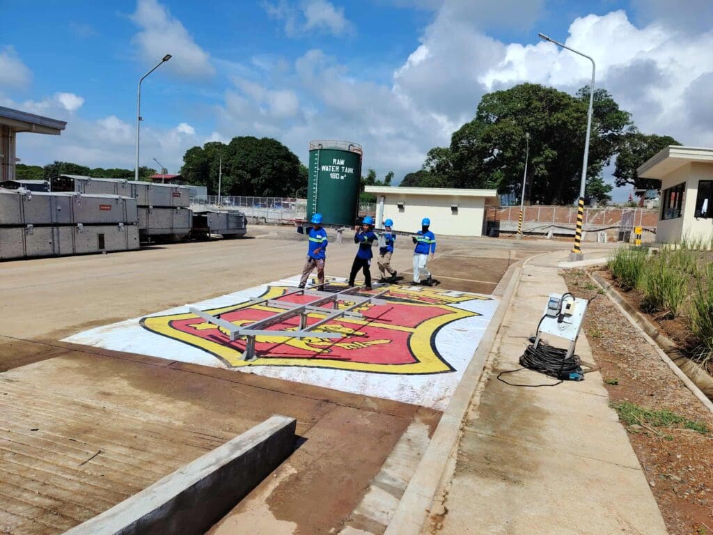 Workers in blue uniforms carry a metal frame over a large painted outdoor crest at an industrial facility, sign maker Philippines by M&G Global Ads.