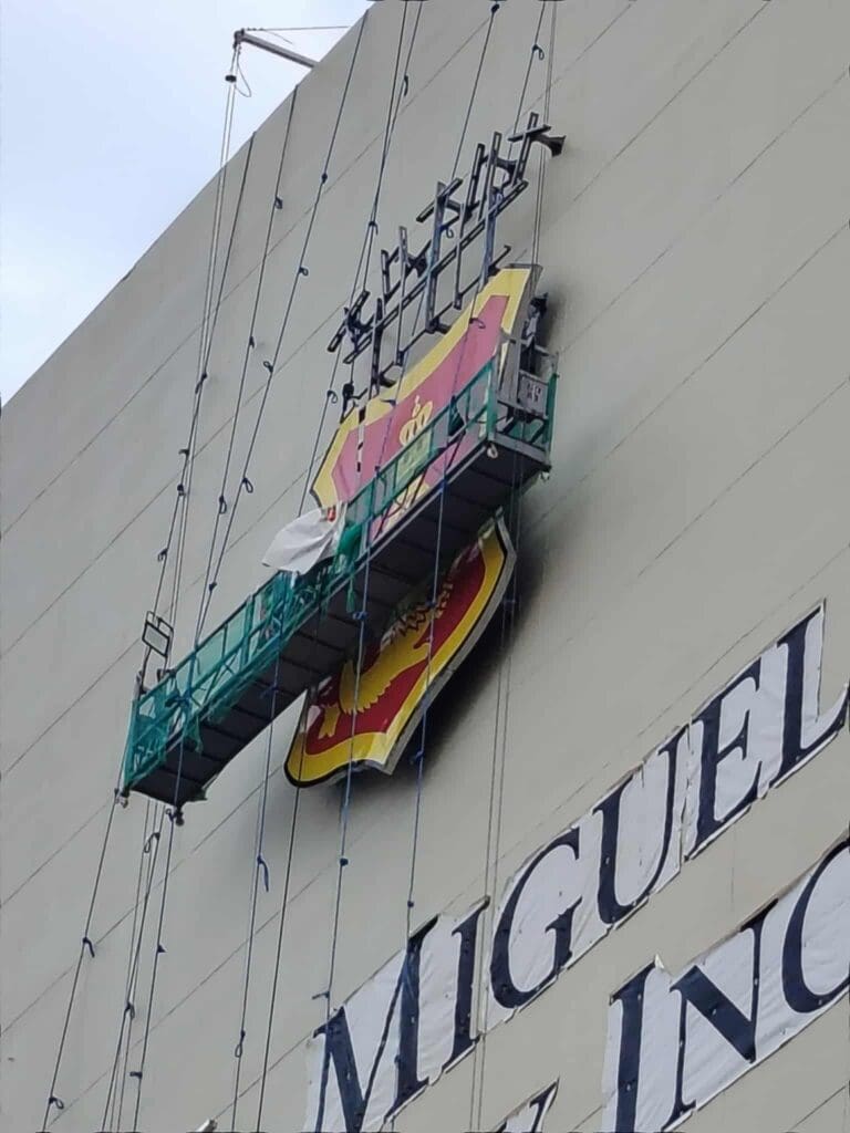 Workers install or repair a large outdoor building acrylic San Miguel logo sign in Metro Manila. Sign maker Philippines by M&G Global Ads.