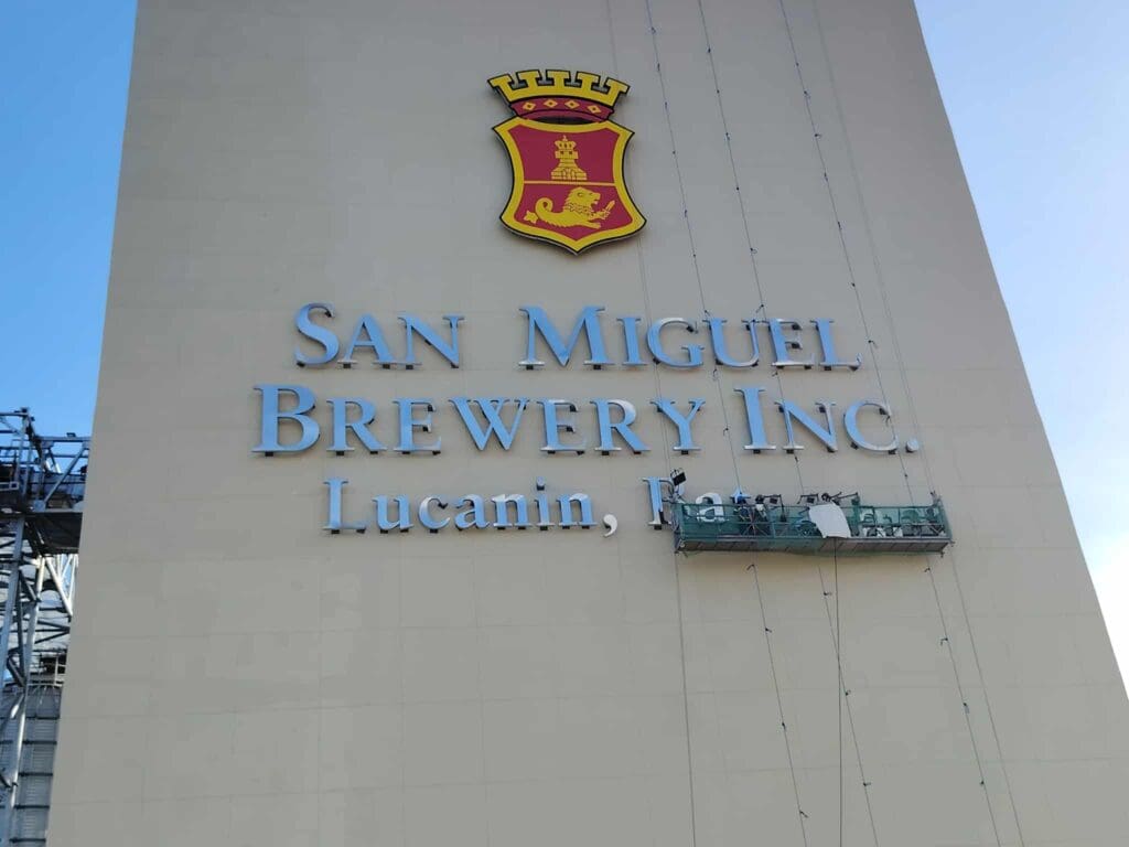 Workers maintain San Miguel Brewery Inc. acrylic building signage with red and yellow crest in Lucanin, Pa.; sign maker Philippines by M&G Global Ads.