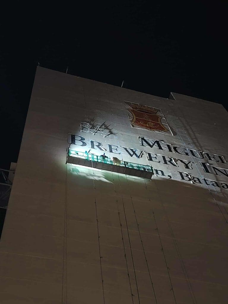 Workers on a suspended platform at night installing building signage for Miguel Brewery, Metro Manila, by sign maker Philippines M&G Global Ads.